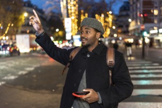 Young man in a winter coat stands on a busy city street at night, holding a smartphone and raising