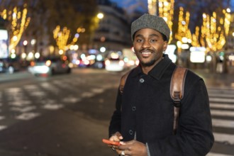 Young black man smiling in an urban setting at night, holding a mobile phone and standing on a city