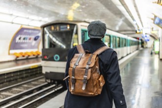 Man with backpack and flat cap standing on a subway platform, watching a train arriving,