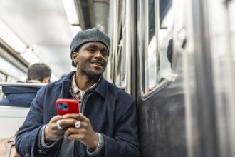 Young black man in beret and jacket rides the subway holding a red smartphone, smiling and looking