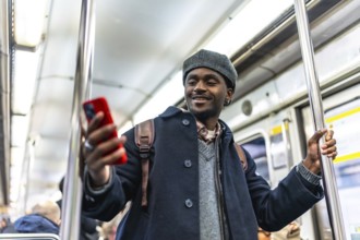 Young black man with a backpack holding onto a pole, smiling while enjoying social media on his red