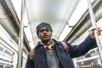 Young black man in winter coat and hat stands holding a subway pole on a crowded metro, looking