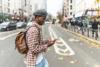 Young man wearing a beret and backpack walking across a city street, looking down at his smartphone