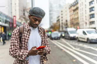 Young black man standing on an urban street, wearing a plaid shirt and a cap, engrossed in using