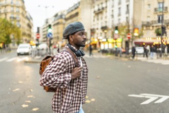Young black man standing on a city street at a crosswalk, wearing a hat and backpack with