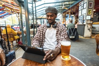 Young black man wearing headphones and flat cap working on digital tablet while sitting at a bar