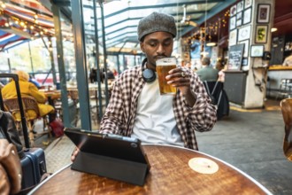 Young man in a plaid shirt and hat relaxing at a pub table, drinking a beer while browsing on a