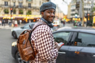 Young black man smiling, wearing a beret and backpack, holding a smartphone while walking on a busy
