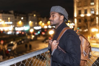Young man in a wool cap and backpack stands on a bridge at night, gazing at the illuminated paris