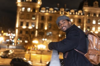 Young man smiling, wearing a beret, coat and backpack, viewing the illuminated city of paris at