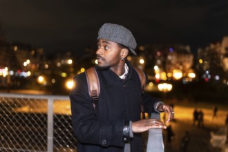 Young man in a flat cap and backpack gazes across a paris bridge at blurred city lights and bokeh