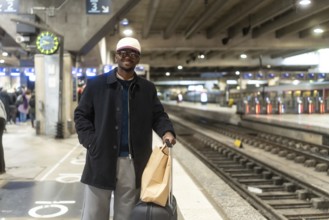 Young black man smiling on a city train platform, wearing a coat and cap, holding luggage and a