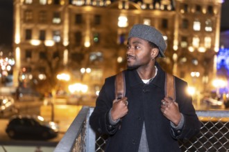Young man in a beret and coat with backpack pauses on an illuminated paris street at night, gazing