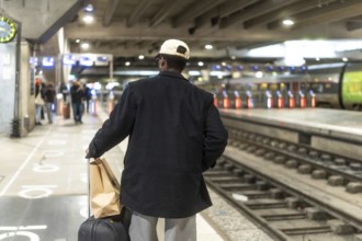 Man standing on a train station platform, seen from behind, pulling a suitcase and carrying a paper