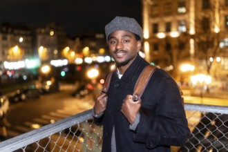 Young african american man smiling, wearing a cap and backpack, standing on a bridge overlooking
