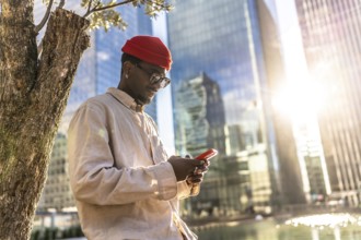 Young black man wearing a red beanie and sunglasses checking his mobile phone outdoors, standing by