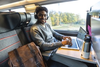 Young african man smiling, wearing headphones and typing on a laptop, working remotely or commuting