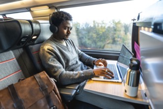 African man traveling on a modern train, wearing headphones and concentrating on his laptop,