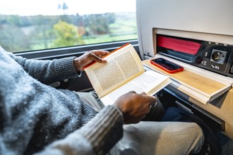 Man reading a book while commuting on a long train journey, using the tray table as a workspace
