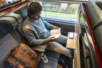 Young professional in headphones sits by a train window reading a book during a high speed