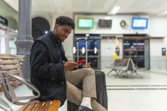 Young black man sitting comfortably on a wooden bench at a train station, using his red smartphone