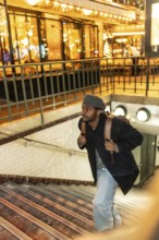 Young black man in beret and coat climbs subway stairs at night, backpack on shoulder, illuminated