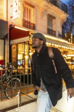 Young black man wearing a beret, coat, and backpack exploring the brightly lit streets of paris,