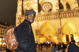 Young black man with a backpack smiling, visiting notre dame cathedral in paris france at night,