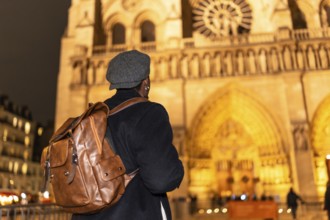Tourist with a backpack and flat cap looking at the iconic notre dame cathedral facade, illuminated