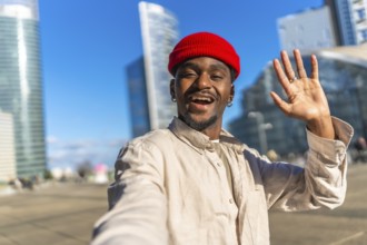 Young black man taking a selfie, smiling broadly and waving at the camera, showing a friendly
