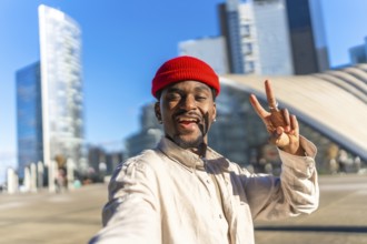 Young black man smiling and making a peace sign gesture while taking a selfie outdoors in a modern