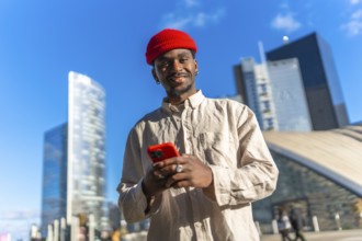 Young black man smiling at camera while using a red smartphone outdoors, standing in a contemporary