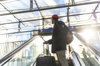 African american man traveling with a suitcase and backpack, riding an escalator upward in a modern
