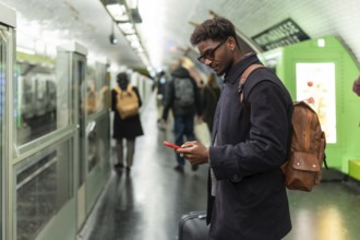Young black man with a backpack and sunglasses standing on a subway platform, checking his