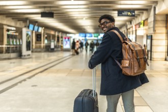 Smiling african man carrying backpack and pulling luggage, looking back while walking through a