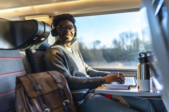 Young black man smiling while traveling on a train, wearing headphones and typing on his laptop,