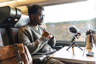 Young black man vlogging on a train, using smartphone and microphone on a tripod during a business