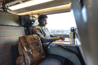 Young man wearing headphones working on a laptop while traveling by train, focusing on remote work