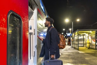 Young man standing on a train station platform at night, holding a smartphone and luggage, looking