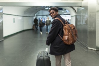 Young man with a stylish leather backpack and rolling luggage is pausing to look back in a modern