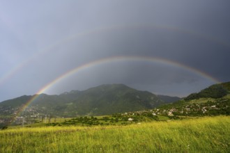 Two rainbows stretch across a green landscape with mountains and a village under a cloudy sky,