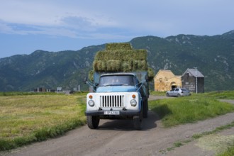 A blue truck with hay bales drives past a church, against a backdrop of mountains, vintage car,