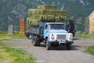 A blue truck with hay bales drives on a dirt road surrounded by mountains and fields, classic car,