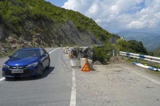 A car drives along a winding mountain road, next to a construction site and lush green areas, road
