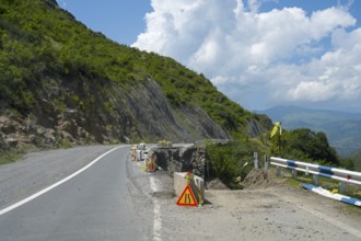 A winding mountain road with a construction site, surrounded by green hills and cloudy sky, road