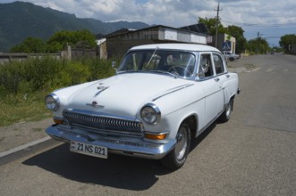 White vintage car on a country road with mountain backdrop and blue sky in summer, GAZ M-21 Volga,
