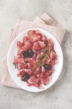 Freshly cut dried meat, served with olives and herbs, on a white plate, light background, no people