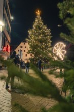 Festively illuminated Christmas tree in a square, people and Ferris wheel in the background,
