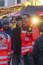 Red Cross paramedics stand at a Christmas market, engaged and friendly amidst the crowd, Aidlingen
