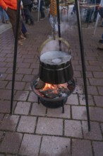 A cauldron hangs over a glowing fire on outdoor cobblestones at a Christmas market, Aidlingen